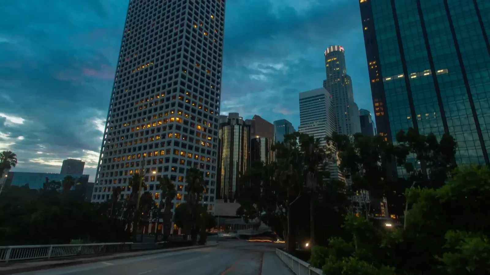 A view of a downtown skyline at twilight. Tall, modern office buildings with lit windows stand against a dark blue sky, viewed from a road with lush foreground trees.